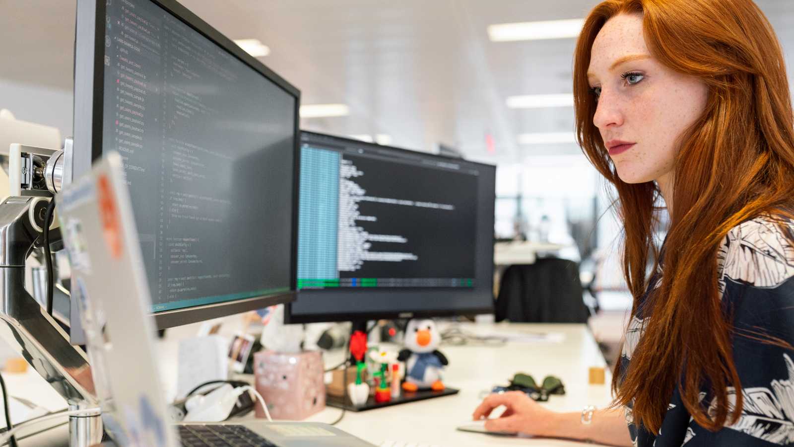 Especialista TI revisando líneas de código en dos monitores desde un puesto de trabajo luminoso